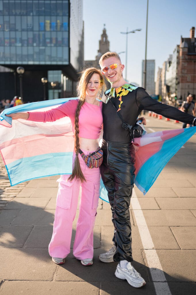 Two individuals posing together outdoors, one holding a transgender pride flag. The person on the left has long hair and wears a pink crop top and baggy pink pants, while the person on the right is wearing a black outfit with colorful accessories and sunglasses.