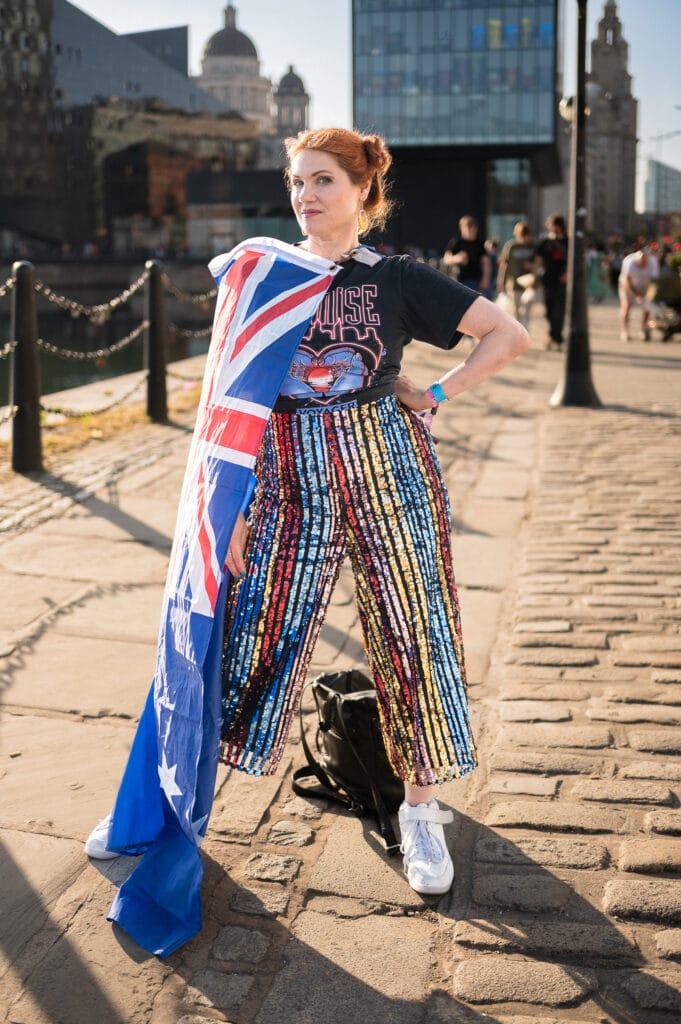 A person standing on a cobblestone path, wearing a black graphic t-shirt and sequin pants, holding an Australian flag draped over their shoulder. In the background, there are buildings and people, with the sun shining.