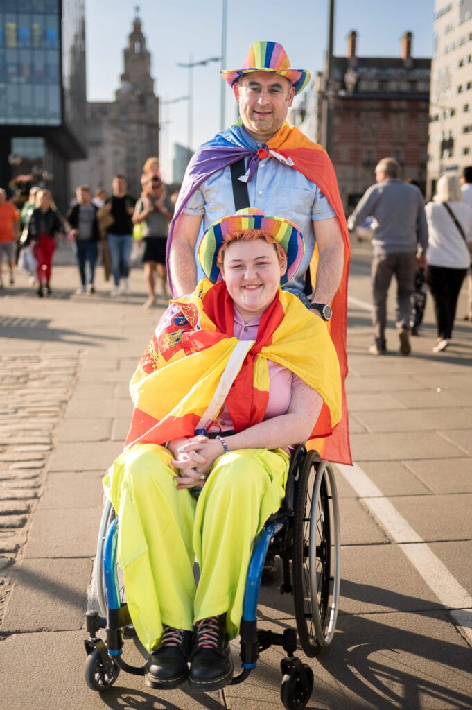 A man and a woman are standing and sitting on a pathway. The man, who is standing behind, wears a rainbow-colored hat and cape. The woman, sitting in a wheelchair in front, also wears a rainbow hat and a colorful outfit. They are surrounded by a crowd of people walking in the background.