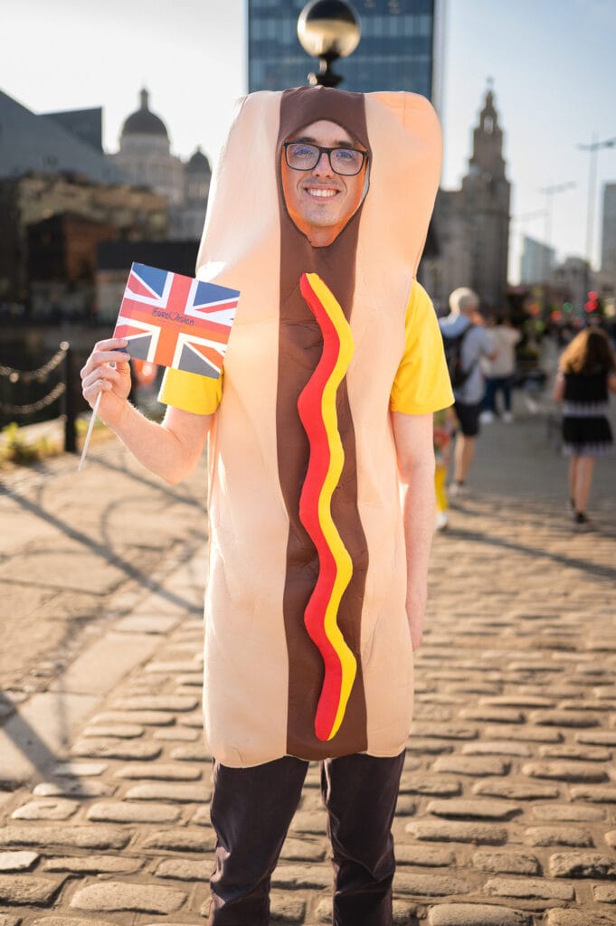A person wearing a hot dog costume stands on a cobblestone path, holding a small British flag. The background includes various buildings and people enjoying the outdoors.