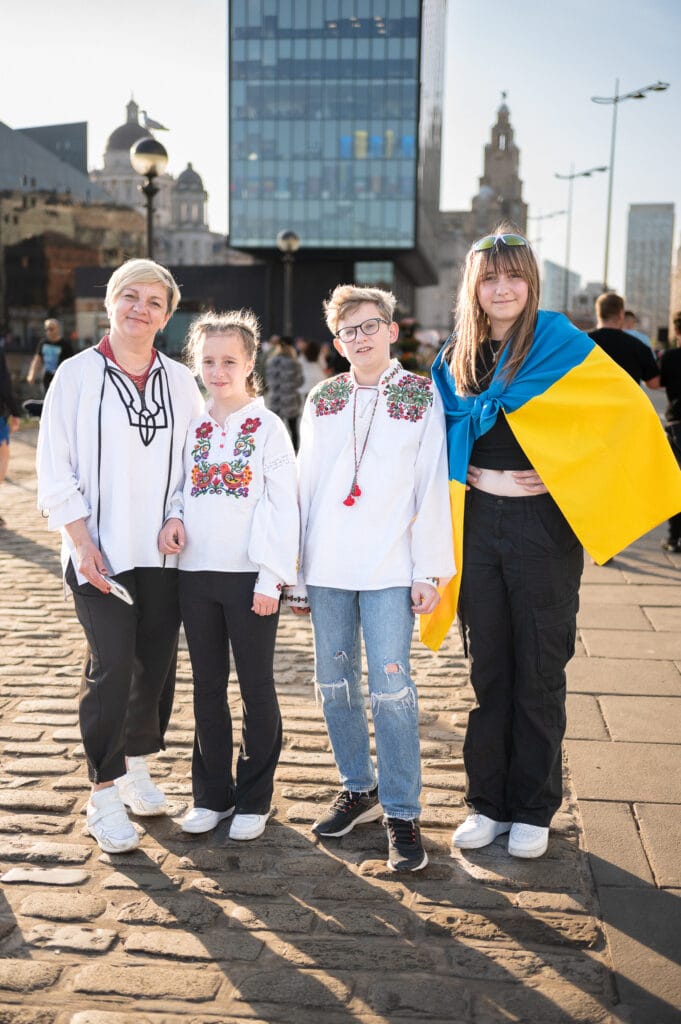 Four individuals standing together outdoors. The first person on the left wears a traditional Ukrainian embroidered shirt, followed by a girl in a similar shirt with floral designs. The third person is a boy wearing glasses and a plain white shirt, and the fourth person on the right is a girl draped in a Ukrainian flag. They are standing on a cobbled surface with buildings in the background.