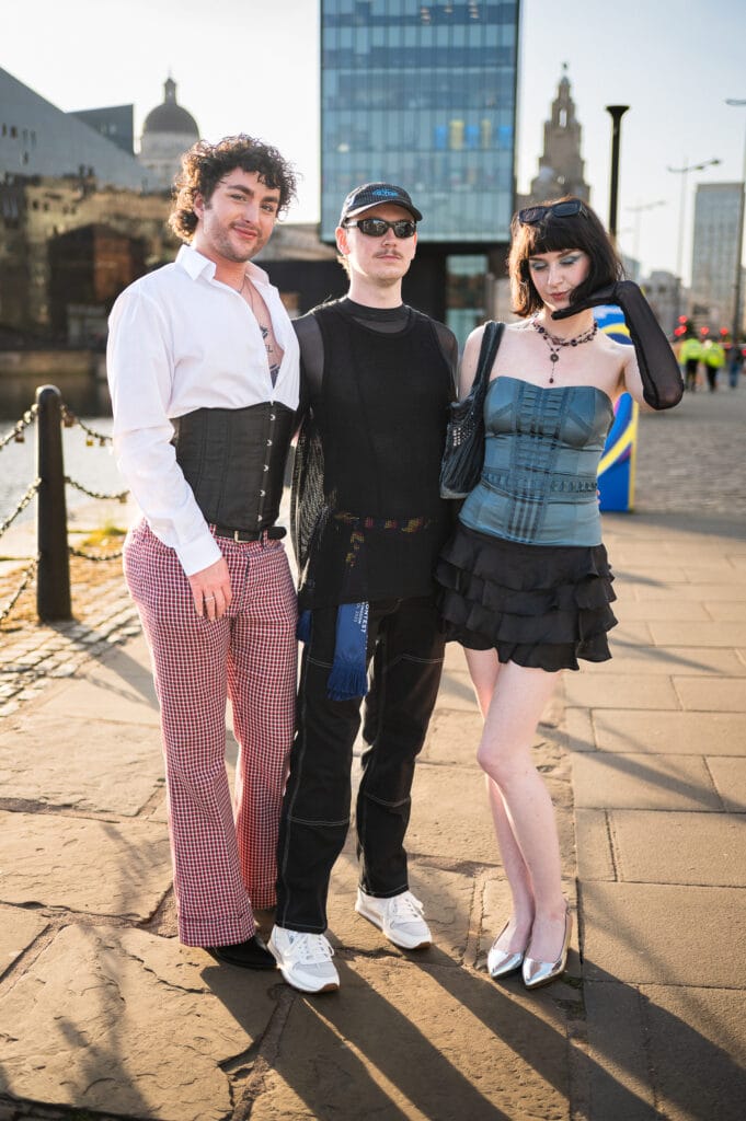 Three individuals posing together outdoors. The first person on the left wears a white shirt with a corset, checkered pants, and has curly hair. The middle person is dressed in a black mesh top and sunglasses. The person on the right is wearing a blue corset top and a black ruffled skirt, with short dark hair. In the background, there are buildings and a waterfront.