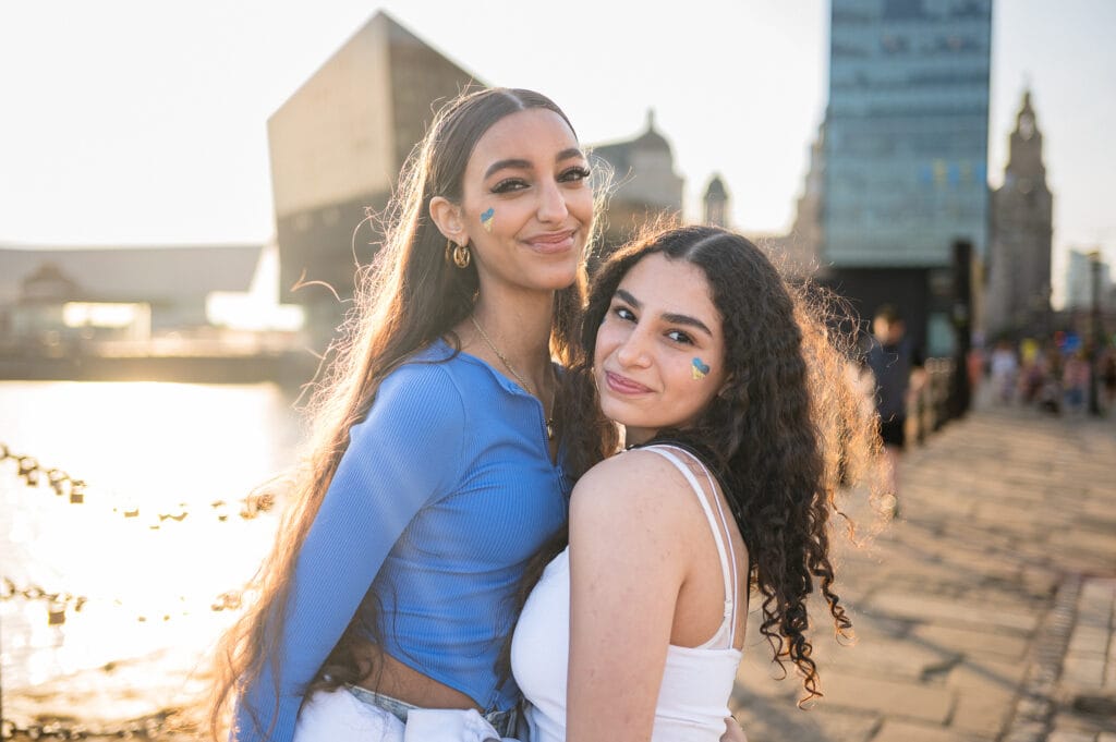 Two women standing close together by a waterfront in an urban area, both smiling. One woman is wearing a blue long-sleeve top, while the other is in a white tank top. Both have face paint in blue and green designs. In the background, there are buildings and a river, with people in the distance.