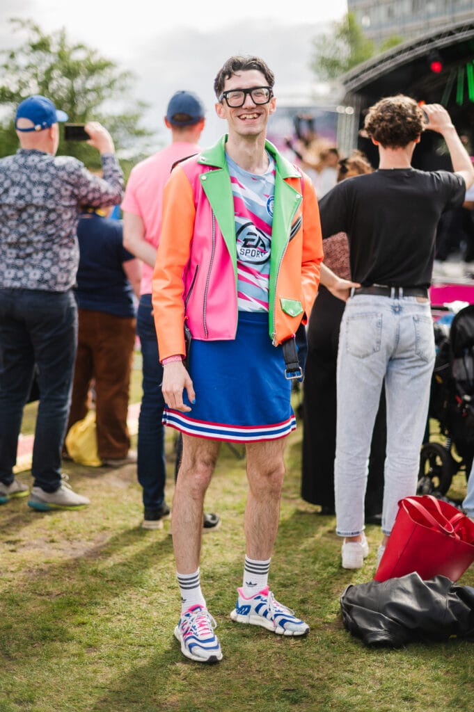 A person smiles and poses in front of a crowd at an outdoor event, wearing a brightly colored jacket, a sporty jersey, a blue skirt, and colorful sneakers with striped socks.