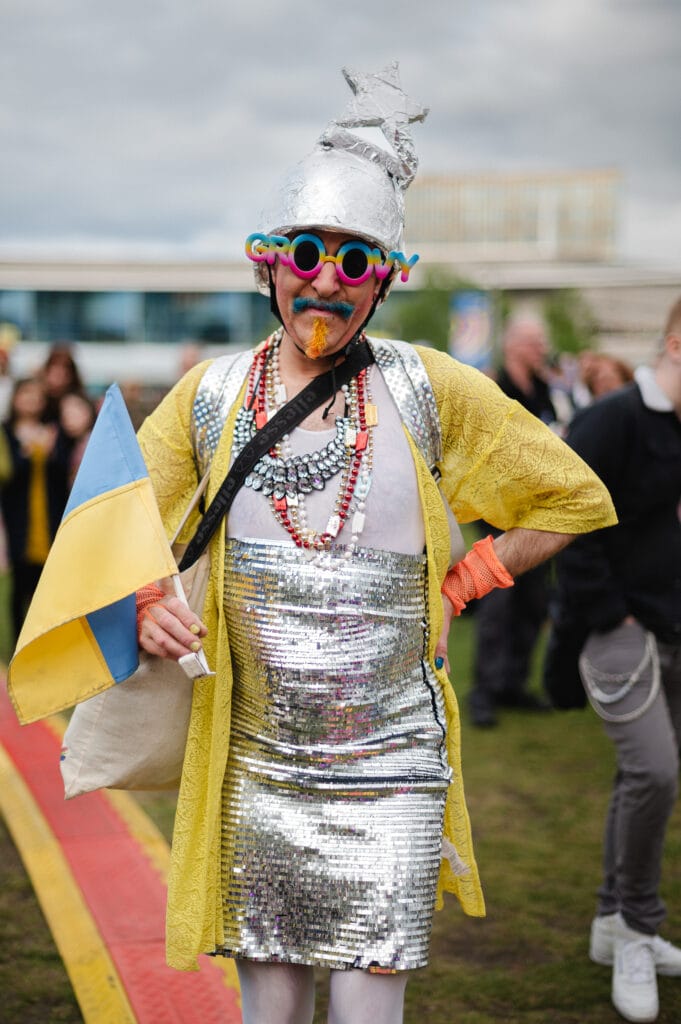 A person dressed in a shiny silver dress with a yellow cardigan, wearing oversized colorful glasses that say "GROOVY," a silver helmet with a star, and orange face paint. They are holding a small Ukraine flag and standing on a grassy area with people in the background.