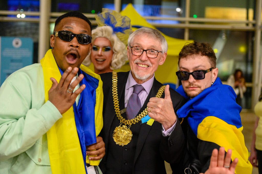 A group of four individuals posing for a photo at an event. Two men in casual outfits are making surprised expressions, while a woman with elaborate makeup and hairstyle stands between them. The man in the center, who is wearing a formal suit and a gold chain of office, is giving a thumbs up. One man is draped in a blue and yellow flag, and there are yellow decorations in the background.