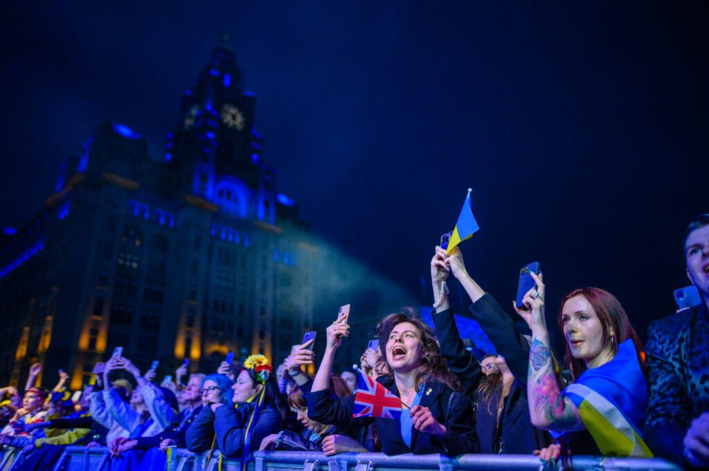 A crowd of people holding flags and mobile phones, with some waving a British flag and others holding Ukrainian flags. They are gathered in front of a large illuminated building at night.