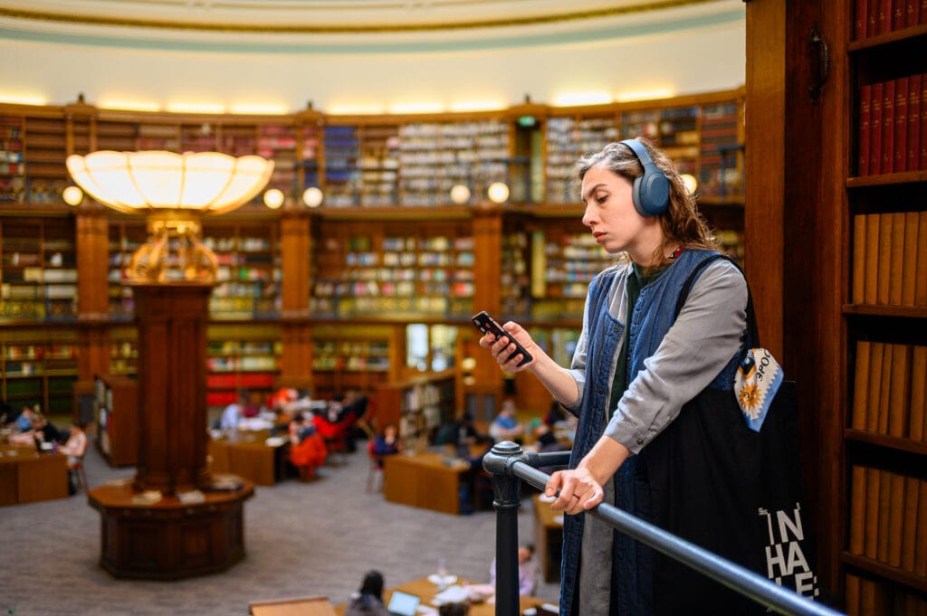 A woman wearing headphones stands on a railing in a library, looking at her smartphone. In the background, shelves filled with books and several people are seated at study tables.
