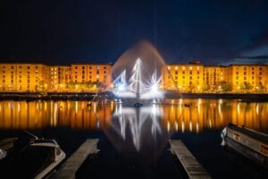 A nighttime view of a waterfront with illuminated buildings reflecting in the water. A fountain sprays water in a decorative arc, and boats are moored at the dock. The buildings, with warm lighting, are brick structures, creating a scenic urban atmosphere.