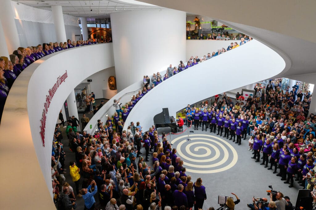A large group of people gathered in an atrium, with many participants wearing purple shirts. They are performing or singing while an audience watches from below and above. A piano is visible in the center, and a spiral design is on the floor.