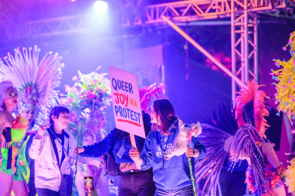 A group of people participating in a vibrant event, holding a sign that says "QUEER JOY is a PROTEST." The individuals are dressed in colorful costumes, with elaborate feathered headdresses and accessories, amidst a lively backdrop of lights and decorations.