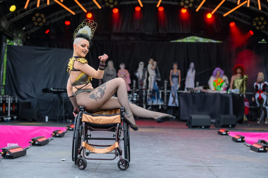 A performer on a stage during a drag show, seated in a wheelchair. The performer has a distinctive outfit with gold accents and a feathered headpiece, one leg extended gracefully. In the background, there are cutouts of other performers and a DJ setup with colorful lighting.