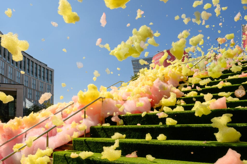 Colorful foam in pink and yellow spills down green steps, with a clear blue sky above and a modern building in the background, suggesting a festive outdoor event in Liverpool.