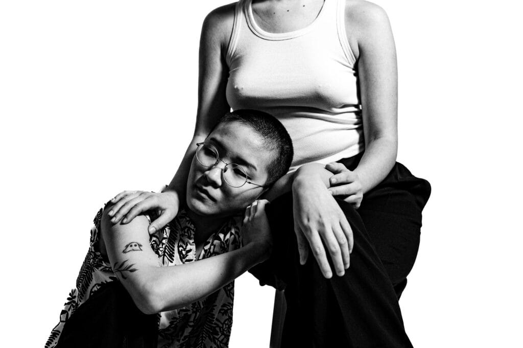 A person with a shaved head and glasses rests their head on the knee of another person seated beside them. The person in the foreground is wearing a patterned shirt, while the seated individual is dressed in a simple tank top. The image is in black and white, conveying a sense of intimacy and connection between the two figures.
