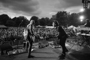A live concert scene featuring two musicians playing guitars in front of a large crowd. The audience is visible, cheering and clapping. The stage is equipped with musical instruments and equipment, and there are trees and tents visible in the background.
