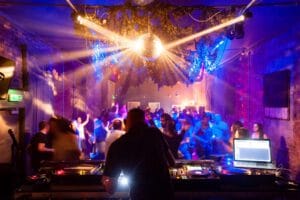 A lively indoor party scene with a DJ in the foreground. The DJ is operating turntables and a laptop, while colorful lights illuminate the space. In the background, people are dancing, some raising their arms, amidst a mix of shadows and light. A disco ball and foliage decorations hang from the ceiling.
