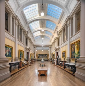 Interior view of the Lady Lever Art Gallery in Port Sunlight, featuring a long, brightly lit hall with high vaulted ceilings, large windows, and ornate columns. The walls are adorned with various paintings in gilded frames, while elegant display tables and sculptures enhance the space.
