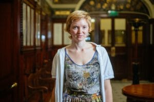 A woman with short, wavy blonde hair is standing in a wooden-paneled room, wearing a floral-patterned top and a light cardigan, looking directly at the camera with a neutral expression.