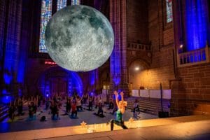 A large moon hangs in a cathedral space, where a group of people are practicing yoga on mats. The interior features stained glass windows and is lit with blue lighting, creating a serene atmosphere. In the foreground, a person is seen in a yoga pose with arms raised.