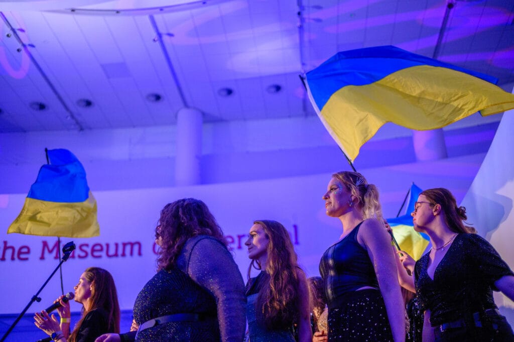 A group of performers on stage holding Ukrainian flags during an event at a museum. The performers are dressed in formal attire, and the backdrop features a sign indicating it's the museum. The venue is illuminated with colored lights.