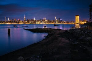A twilight view of the New York City skyline from a waterfront, featuring illuminated skyscrapers including the Empire State Building, with two people standing on a rocky shore in the foreground.