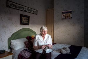 An elderly woman with short, grey hair is knitting while sitting on a bed with a green headboard. The room is softly lit and features floral wallpaper, a picture of a cow, and a small stuffed bear on the bed.