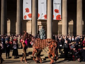 A large, puppet-like horse figure is being led by a performer in front of a gathering of people at a commemorative event. Banners with the phrases "We will remember" and poppy designs hang behind a statue. The crowd includes military personnel and civilians, many wearing red poppies.