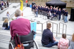 A group of men in dark blue sweaters performing on an outdoor stage, with microphones set up. Some audience members are seated on steps, including a person in a wheelchair and a child. In the background, there is a black tent and various spectators watching the performance.