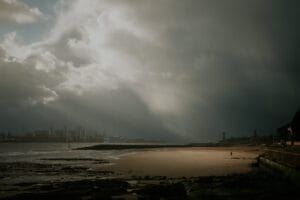 The image depicts a sweeping coastal landscape with a dramatic sky overhead. The beach in the foreground is large and sandy, with rocky outcroppings visible along the shoreline. In the distance, the skyline of a city can be seen, suggesting this is an urban coastal setting. The clouds above are dark and swirling, creating an ominous yet captivating atmosphere. The overall composition and lighting give the scene a moody, almost apocalyptic feel, with the contrast between the expansive beach and the looming cityscape adding to the sense of scale and isolation.​​​​​​​​​​​​​​​​