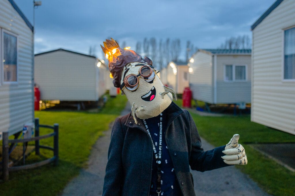 A person wearing a quirky mask and costume, resembling a cartoonish character with a crown of lights, is giving a thumbs-up gesture. The background features several mobile homes and a grassy area, with an evening sky above.