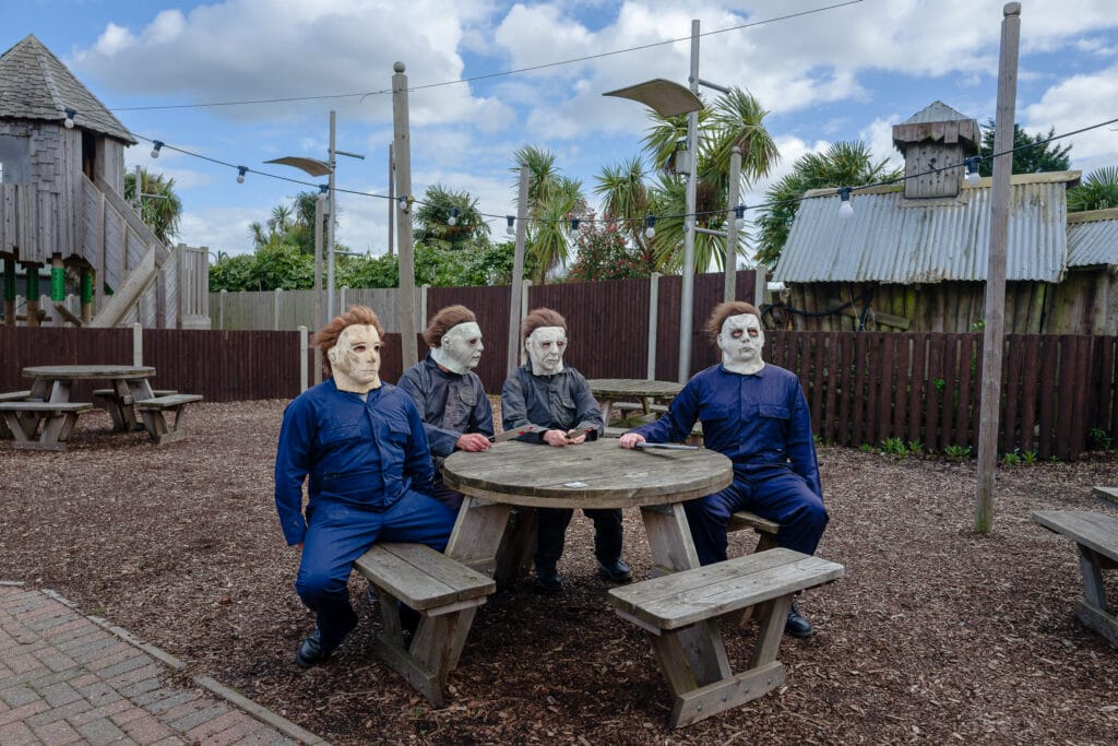 Four figures wearing similar horror movie masks and blue clothing are seated around a circular wooden table in a playground area. In the background, there are playground structures and palm trees under a partly cloudy sky.