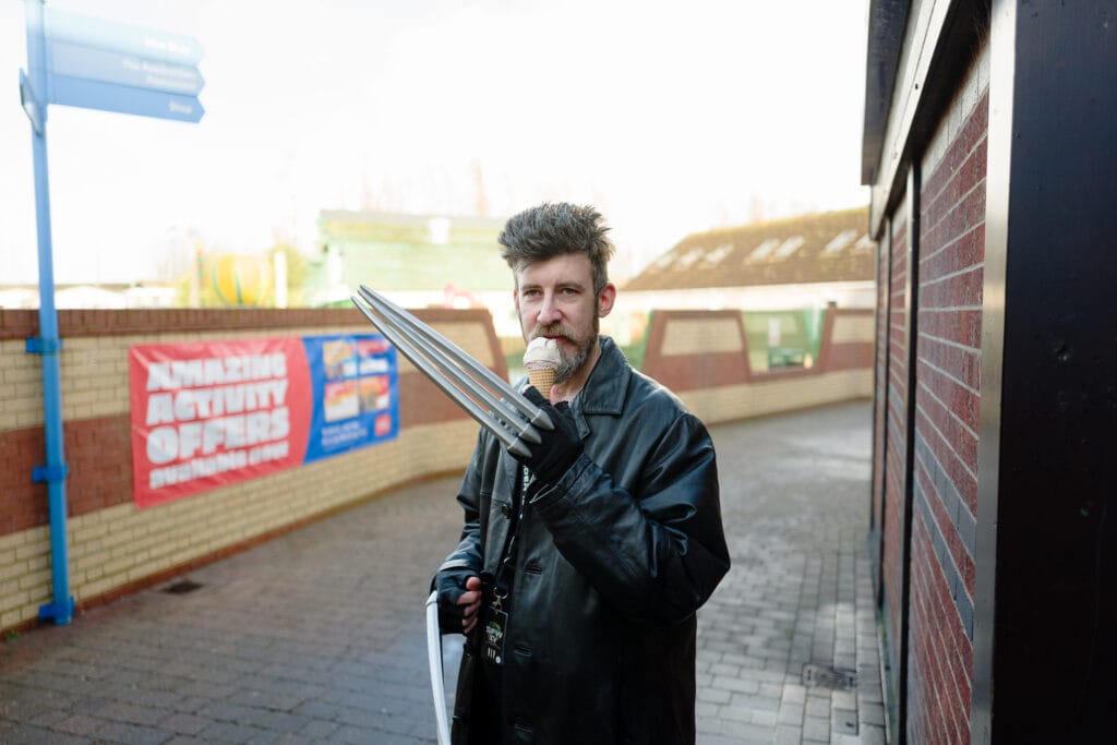 A person wearing a black leather jacket is holding a cone of ice cream in one hand and a set of claw-like props in the other hand. They are standing in a pedestrian area with a colorful background including a banner that says "Amazing Activity Offers."