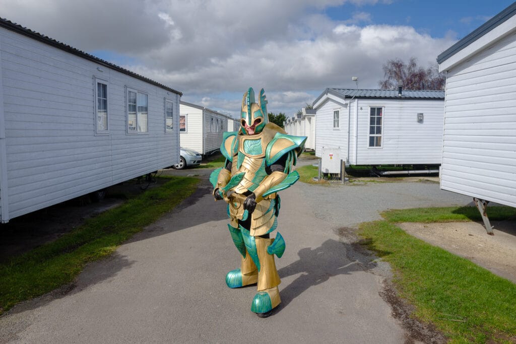 A person dressed in a colorful, armored costume resembling a fictional character stands on a path between two rows of white mobile homes. The scene is set outdoors with a partly cloudy sky.