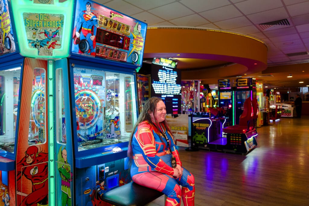 An individual wearing a colorful superhero costume sits on a stool in an arcade. Behind them, there are various arcade games, including a Superman-themed game and a racing game, illuminated with vibrant lights.