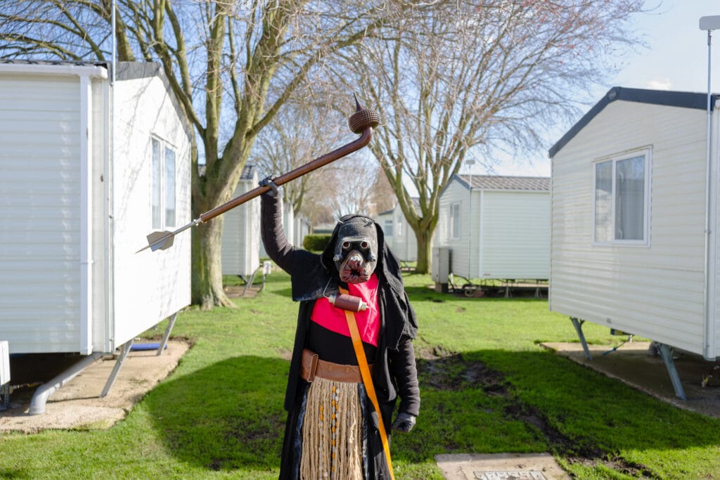 An individual dressed in a costume resembling a hooded figure, holding a staff with a small object on top, stands in a grassy area between two white mobile homes. The person has a dog mask on, and the outfit features black robes and a red and orange costume underneath. Trees can be seen in the background, and it appears to be a sunny day.