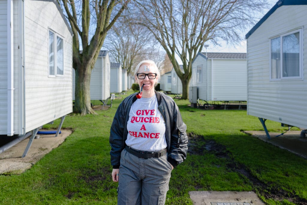 A person standing in a grassy area between two mobile homes, wearing a grey shirt with red text that says "GIVE QUICHE A CHANCE" and grey pants. The person has glasses, a light-colored hat, and is holding a cigarette in their mouth.