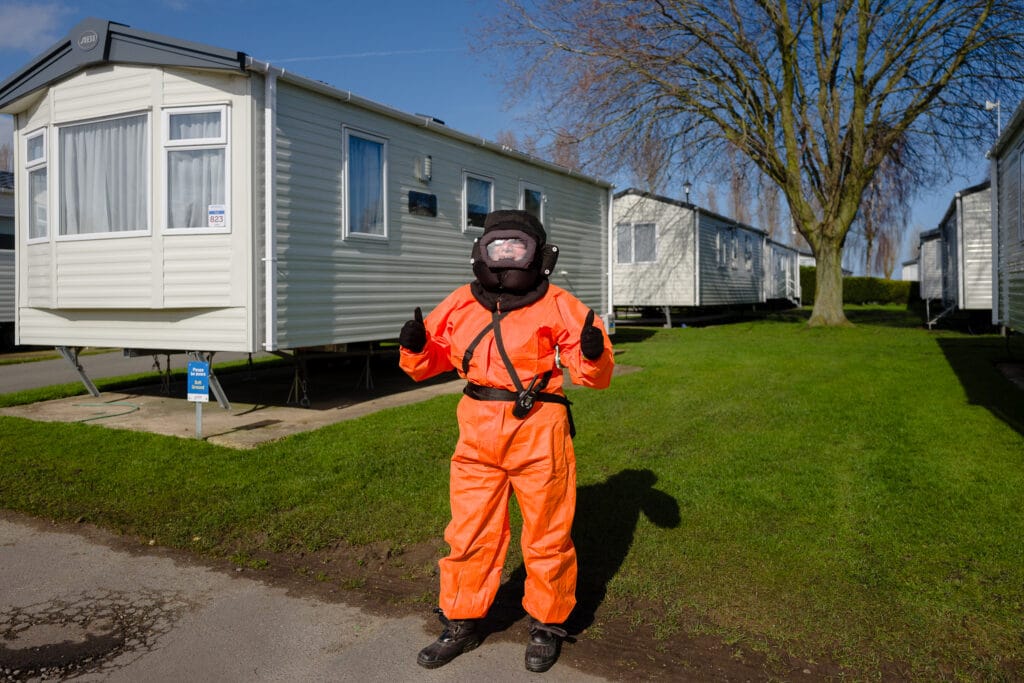 A person dressed in an orange protective suit and helmet is standing in front of a row of mobile homes, giving a thumbs up. The scene features green grass and a bare tree in the background.