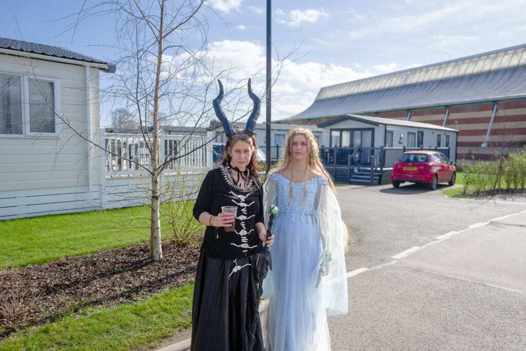 Two women are standing outside near a building. One woman is wearing a black dress with horn-like accessories and holding a drink, while the other is wearing a light blue gown and holding a flower. In the background, there are additional buildings and a parked red car.