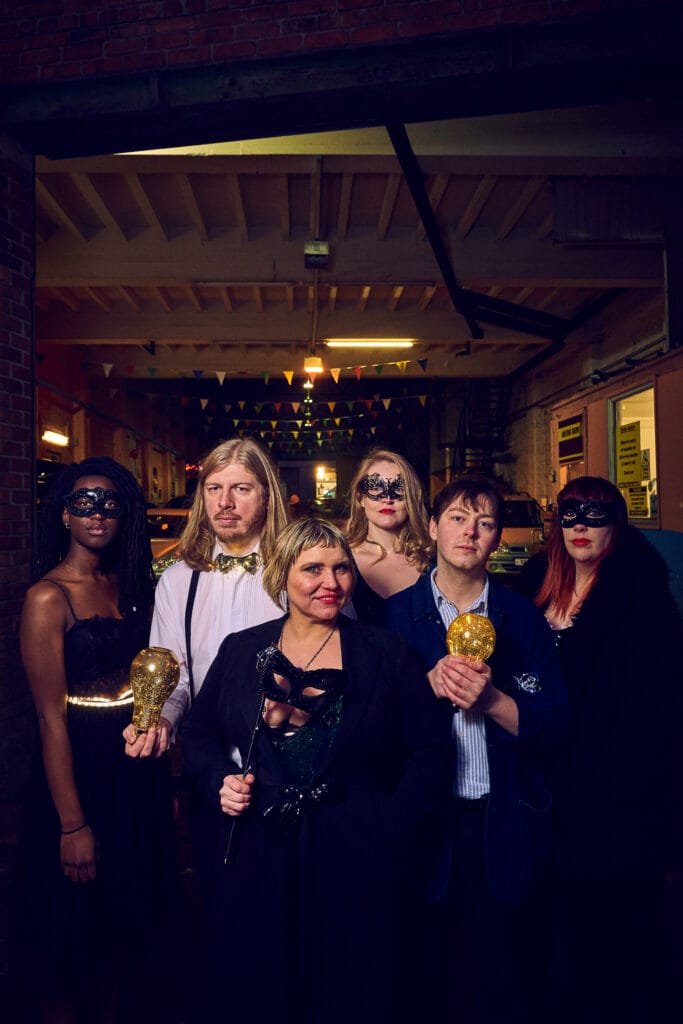 A group of six people posing together in a dimly lit venue. They are dressed in formal and semi-formal attire, with some wearing masks. A woman in the center holds a stick, while others are holding golden objects. Colorful bunting hangs in the background.