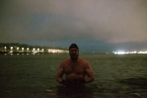 A shirtless man standing in water at night, wearing gloves and a swim cap. A cityscape with lights is visible in the background along a shoreline.