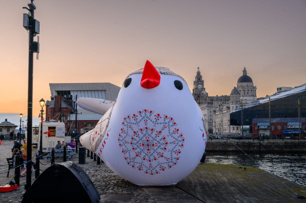 A large inflatable white bird with a red beak and patterned details is situated in a harbor area. In the background, historic buildings and a modern structure are visible along the waterfront, with people nearby and a streetlamp illuminating the scene.