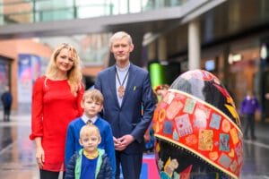 A group of four people stands together in a modern indoor space. A woman in a red dress is on the left, next to a man in a dark suit and a white shirt, who is wearing a large cross necklace. In front of them are two boys, one wearing a blue sweater with a white collar and the other with blonde hair in a green jacket. Next to them is a large decorated egg sculpture featuring various colorful designs.