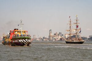 A colorful tugboat and a tall ship sailing on the water, with a city skyline in the background. The tugboat is painted in vibrant colors and patterns, while the tall ship has multiple sails and flags. The scene captures a clear day with distant buildings visible behind the boats.