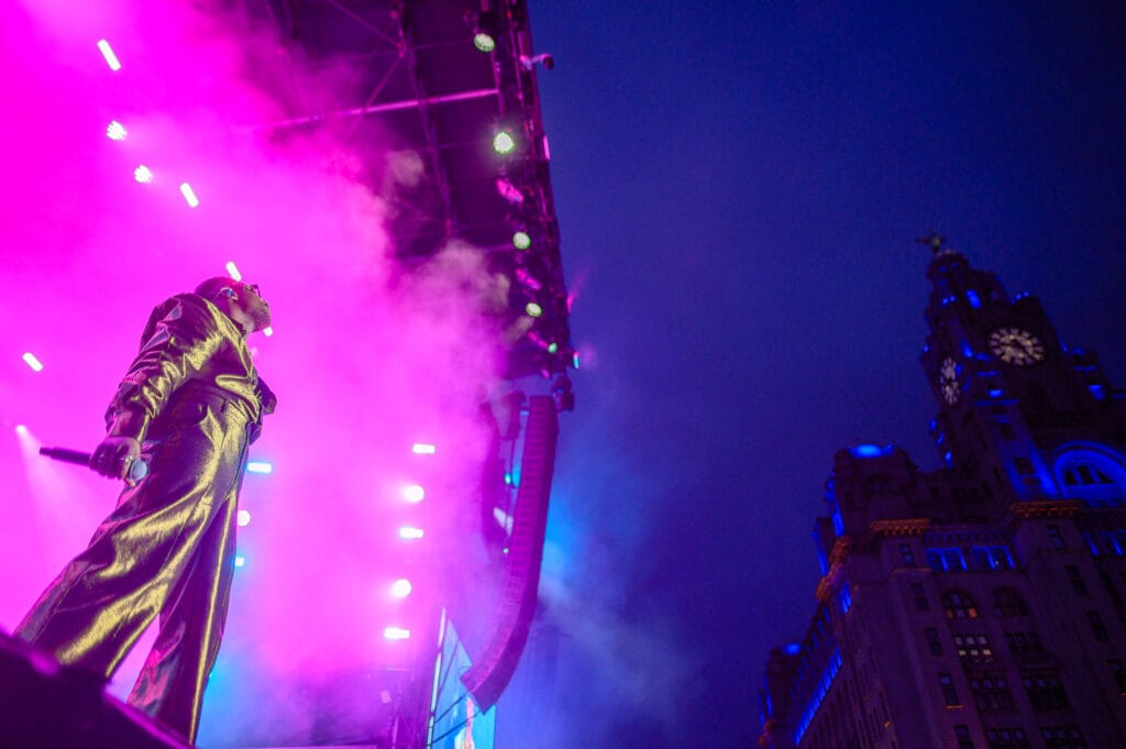 A performer on stage wearing a shiny gold outfit, holding a microphone, is illuminated by colorful stage lights and smoke. In the background, a historic building with a clock tower is visible against a twilight sky.