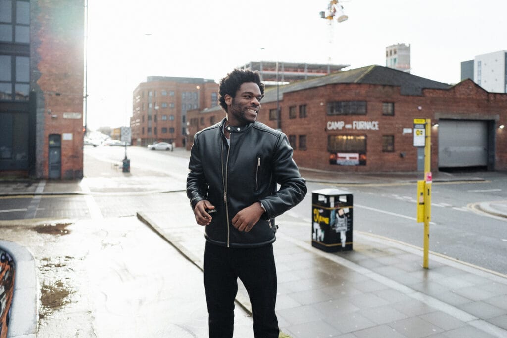 A man wearing a black leather jacket stands on a city street. He is smiling and walking towards the camera. In the background, there are red brick buildings and modern structures, along with a street sign and a trash bin.