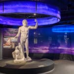 A large white statue of a muscular male figure stands on a circular pedestal within an exhibition space. Above the statue, an illuminated purple circular feature enhances the ambiance. In the background, colourful displays and posters provide context about the exhibition, while artifacts are showcased in glass cases to the left.