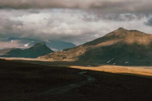 A scenic view of rugged mountains under a cloudy sky in Iceland, featuring a winding dirt road and areas of light and shadow on the landscape.