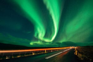 A night scene featuring the northern lights in vibrant green hues swirling across the sky, with a dark road illuminated by moving vehicle lights and distant power lines visible in the background.
