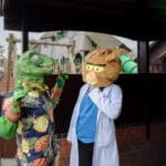 Two people in cosplay, one dressed as a dinosaur and the other as a cat, stand talking near a playground.