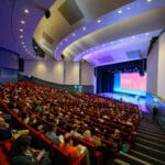 A large audience sits in red seats in an auditorium listening to a speaker on stage. The stage has a large screen displaying the text "Introduction: Tom Cassidy."